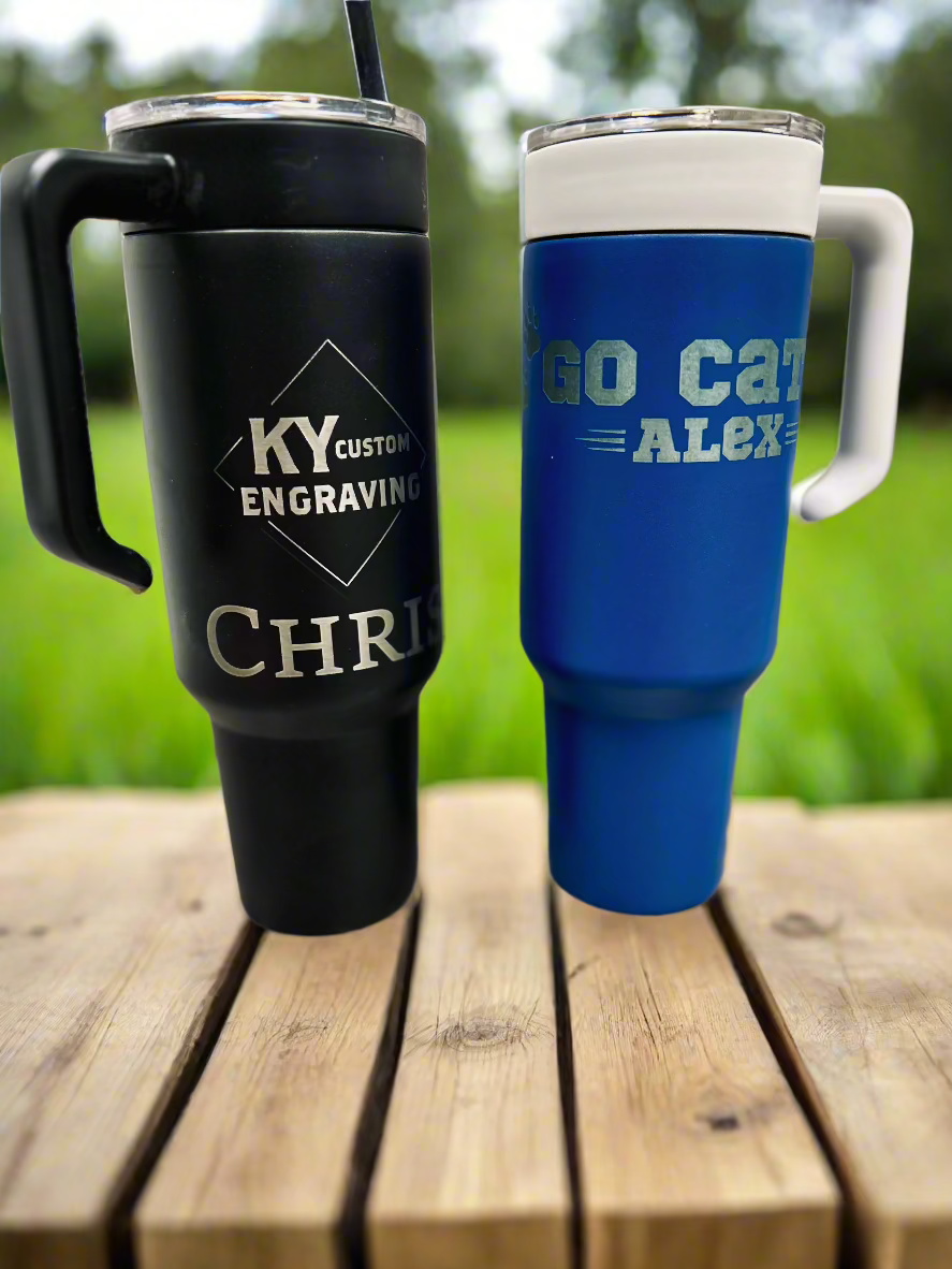 Two custom-engraved travel tumblers with handles placed on a rustic wooden surface outdoors. The black tumbler features 'KY Custom Engraving' and the name 'Chris,' while the blue tumbler displays 'Go Cats' and 'Alex.' Both insulated tumblers have clear lids, making them perfect for hot or cold drinks on the go.