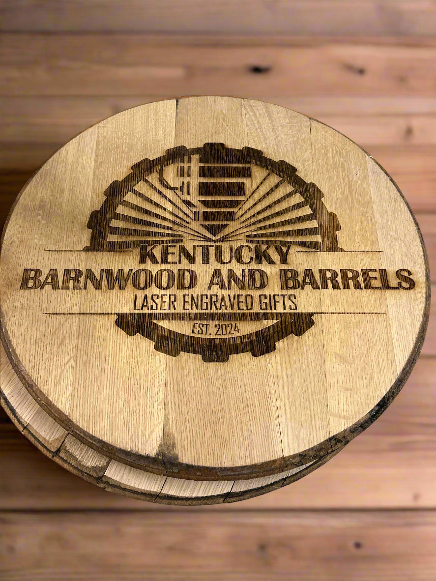 A wooden barrel lid with a laser-engraved logo for "Kentucky Barnwood and Barrels." The design features a rustic barn illustration with sun rays behind it, encircled by a gear-like border. The text "Kentucky Barnwood and Barrels" is prominently displayed, followed by "Laser Engraved Gifts" and "Est. 2024" at the bottom. The lid rests on another wooden barrel, with a warm-toned wooden floor in the background.