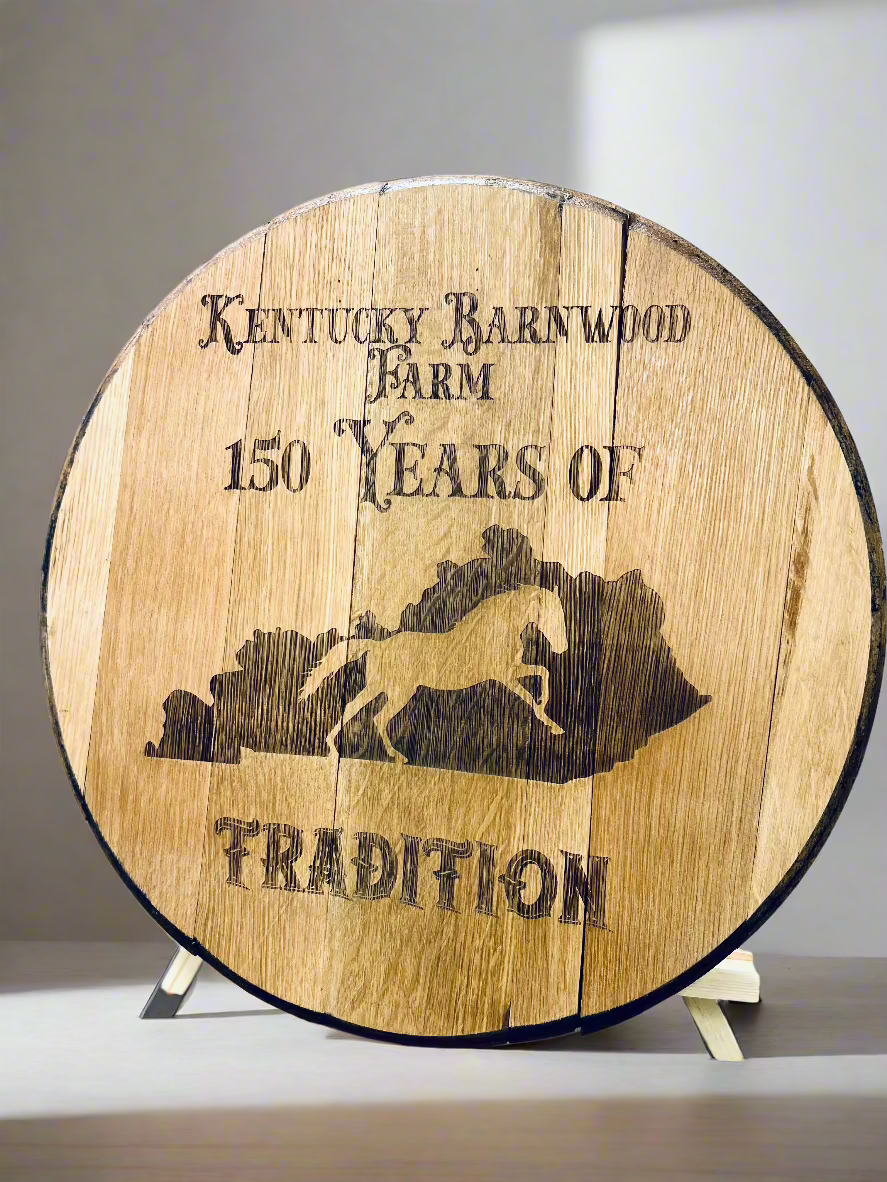 A rustic wooden barrel lid with a laser-engraved design celebrating "Kentucky Barnwood Farm" and "150 Years of Tradition." The engraving features a silhouette of a galloping horse overlaid on an outline of the state of Kentucky. The wood has a natural, aged appearance, with visible cracks and grain patterns, and is displayed on a stand against a neutral background.