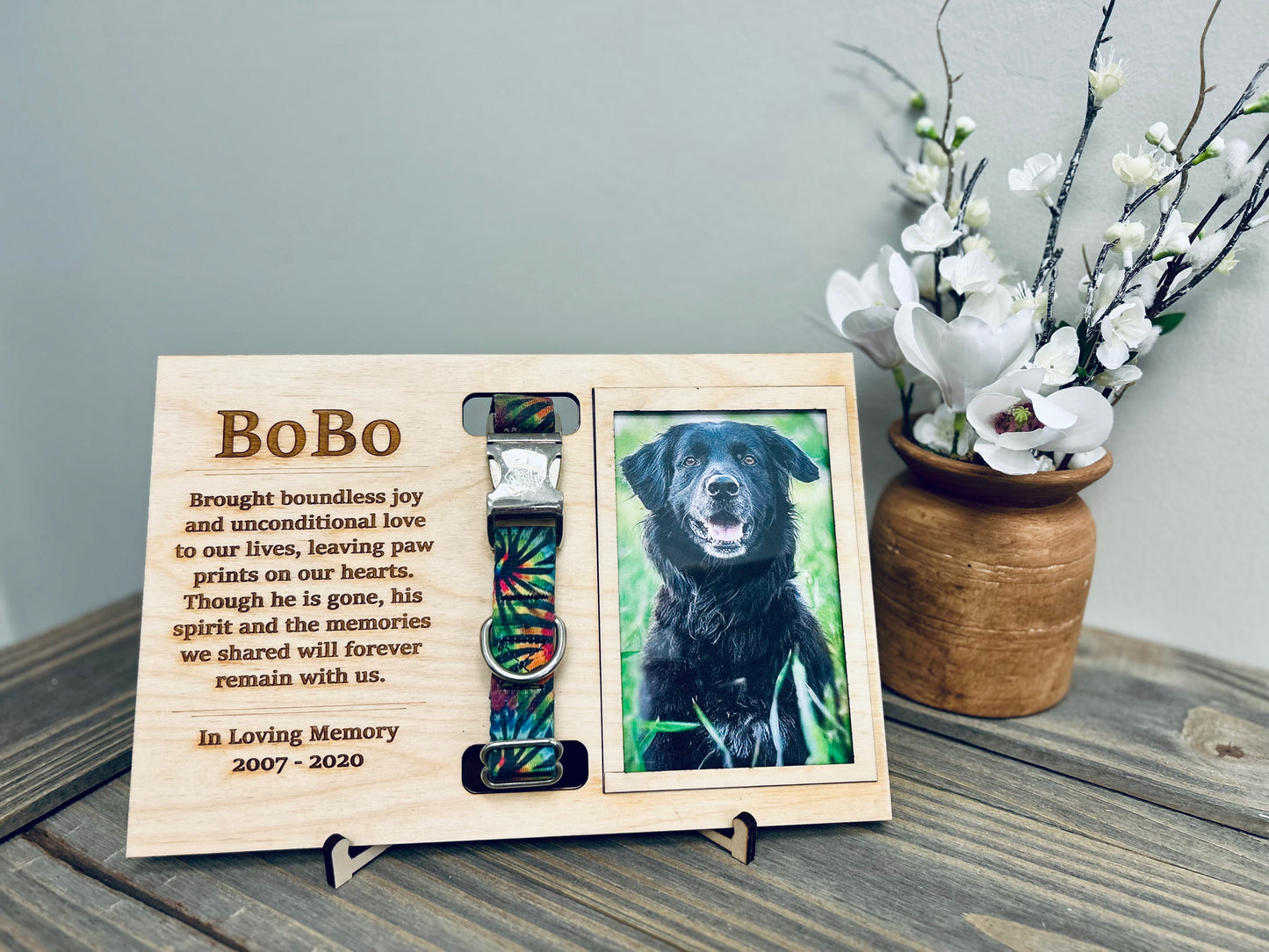 A custom wooden pet memorial plaque dedicated to "BoBo," featuring an engraved heartfelt message about the dog's unconditional love and cherished memories. The plaque includes a slot holding BoBo's colorful collar and a framed photograph of BoBo, a black dog with a joyful expression, sitting in the grass.