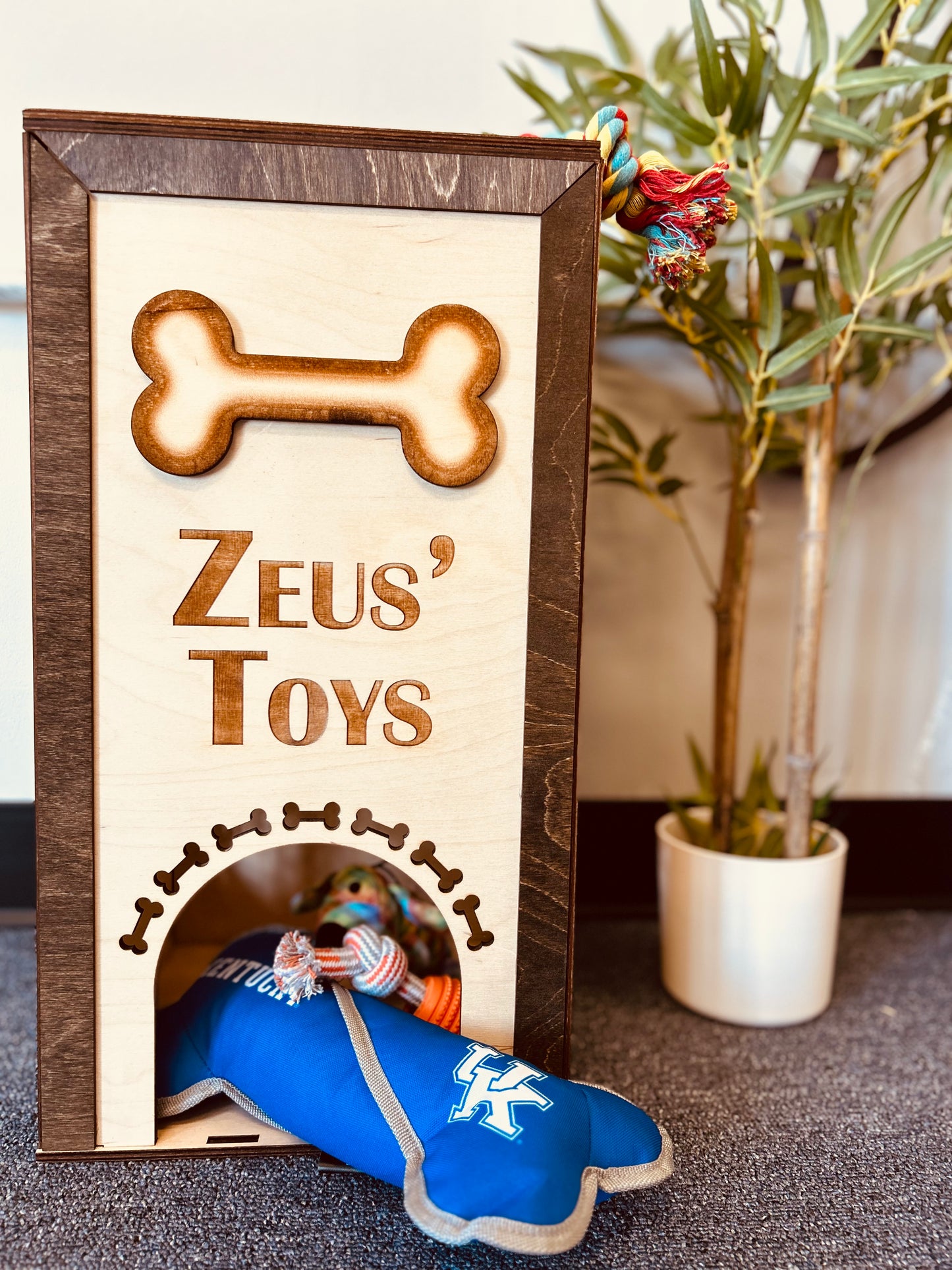 A custom wooden dog toy box engraved with "Zeus' Toys" and a large bone design at the top. The front panel has an arched cutout decorated with engraved bones, allowing easy access to the toys inside. Various dog toys, including a blue plush toy with a University of Kentucky logo and a colorful rope toy, are spilling out of the box. The box is placed on a carpeted floor near a potted plant, adding a cozy touch to the scene.