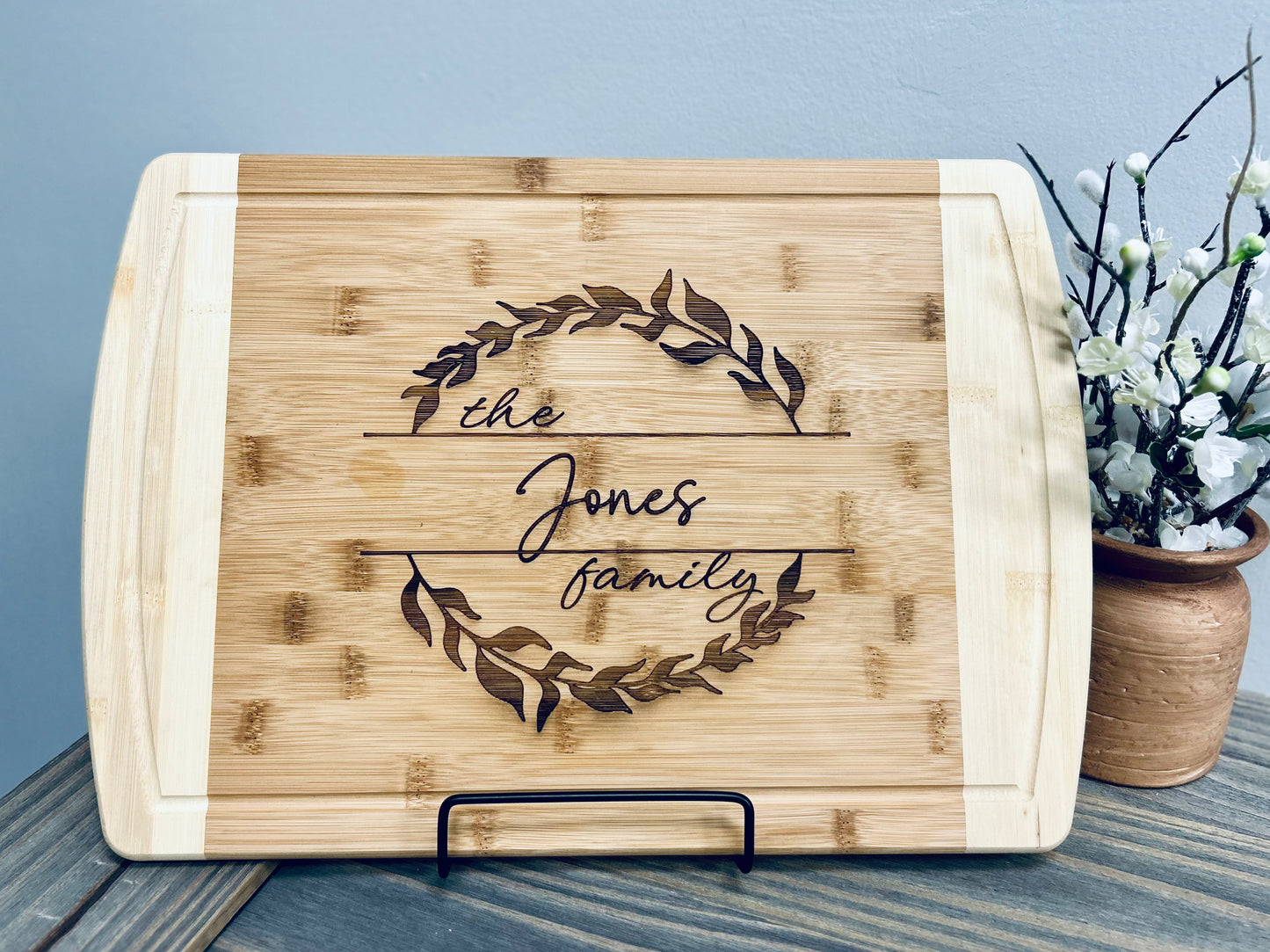 A personalized wooden cutting board engraved with "The Jones Family" in elegant script, surrounded by a delicate laurel wreath design. The board is made of light bamboo with darker engraved details, giving it a rustic yet sophisticated look. It is displayed on a black metal stand on a wooden surface, accompanied by a ceramic vase with white flowers, adding a decorative touch. The engraving makes it a thoughtful and stylish kitchen or home décor piece.