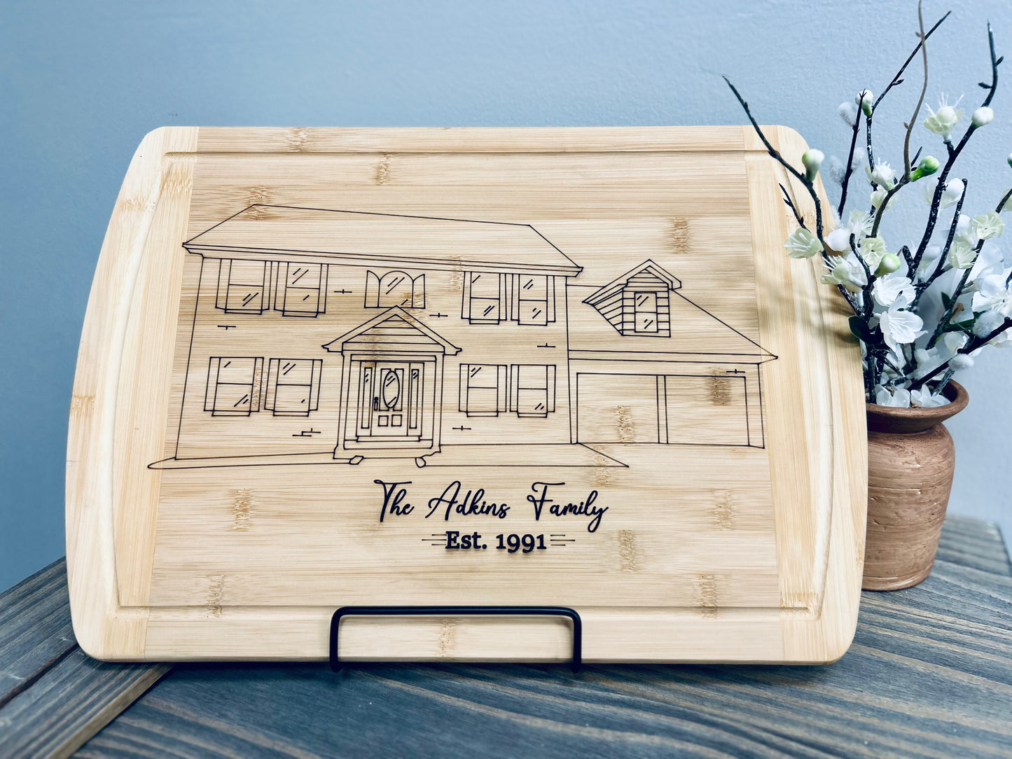 A custom bamboo cutting board featuring a detailed laser-engraved illustration of a two-story family home with a garage. Below the house illustration, "The Adkins Family" is elegantly engraved, followed by "Est. 1991." The board is displayed on a black metal stand and placed on a rustic wooden surface. A vase with delicate white flowers adds a charming decorative touch to the background.