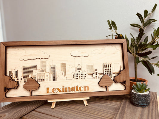 A beautifully crafted wooden artwork featuring a laser-cut skyline of Lexington, Kentucky. The piece is framed with a darker wood border and includes intricate details of the city's buildings, trees, and clouds. The word "Lexington" is prominently displayed at the bottom in a bold, cut-out font. The artwork is placed on a wooden stand and accompanied by potted plants, creating a warm and natural aesthetic.