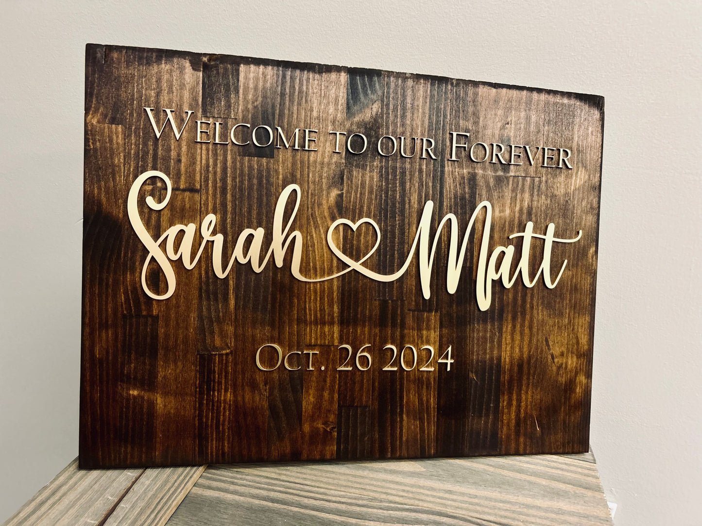 A beautifully crafted wooden wedding sign with a dark-stained finish, featuring laser-engraved and cut lettering. The sign reads, "Welcome to our Forever," followed by "Sarah ♥ Matt" in an elegant script font, and "Oct. 26 2024" below. The names have a heart symbol connecting them, adding a romantic touch. The wood grain is visible, and the sign is displayed on a wooden surface against a neutral wall.
