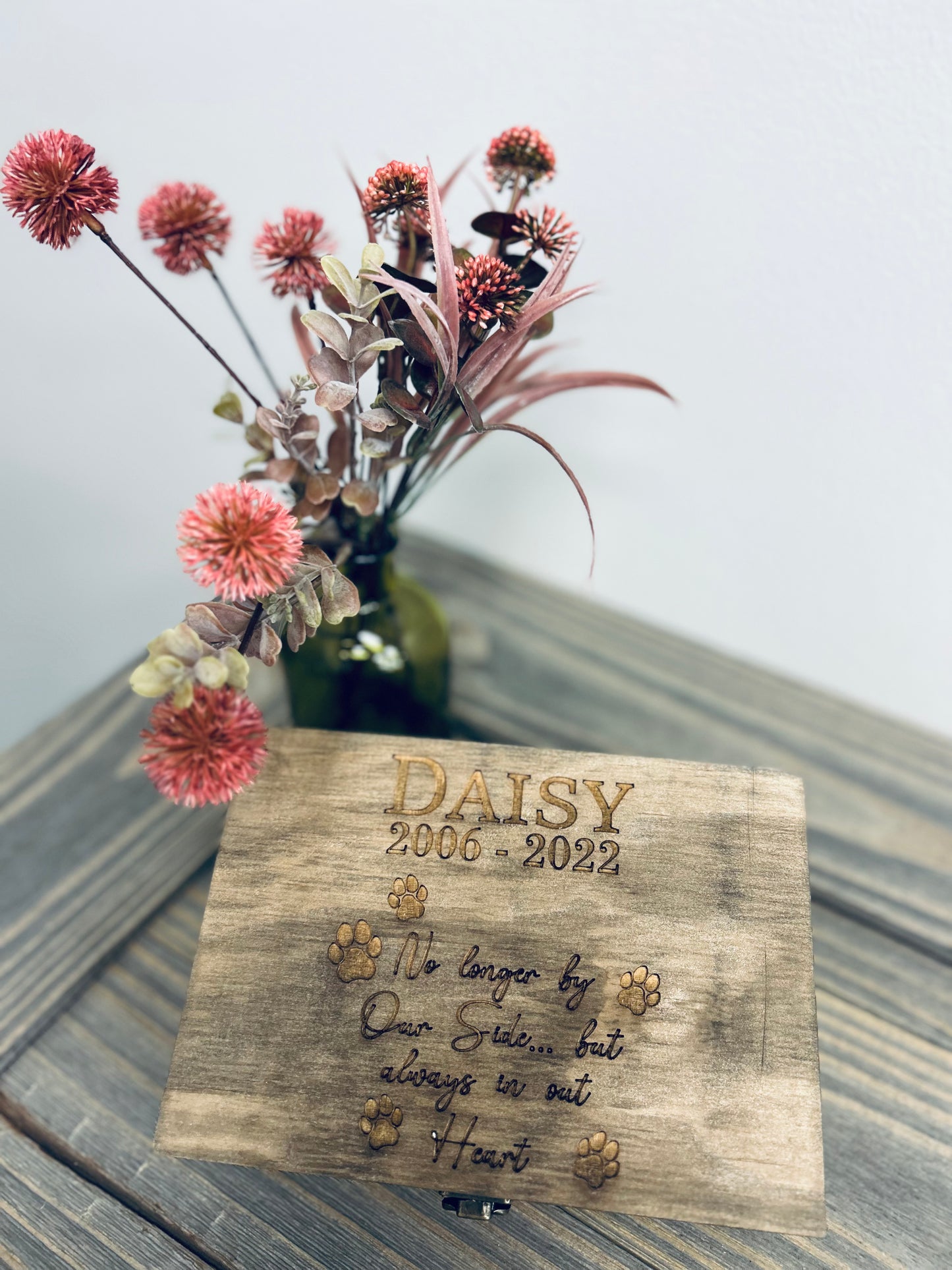 A rustic wooden pet memorial plaque dedicated to "DAISY" (2000-2022), featuring engraved text and paw print designs. The inscription reads, "No longer by Our Sides... but always in our Heart." The plaque is displayed on a wooden surface with a small stand, accompanied by a vase of red and pink artificial flowers, creating a heartfelt tribute in memory of a beloved pet.