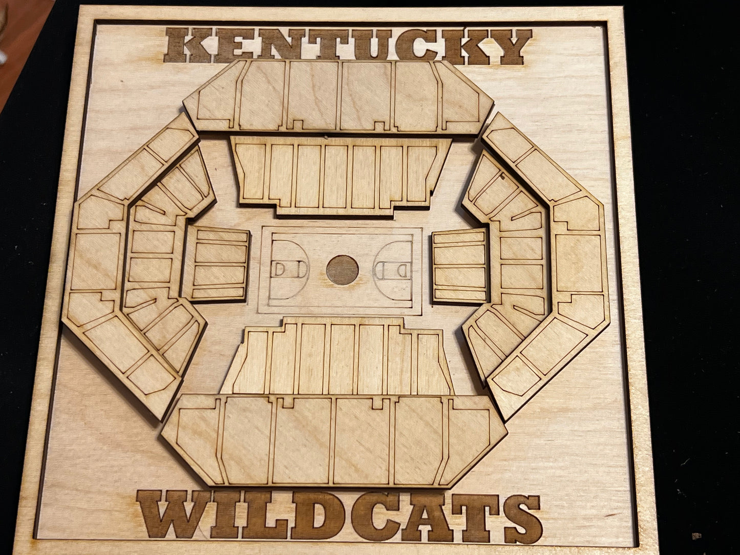 A wooden, laser-cut 3D representation of a basketball arena, labeled "KENTUCKY WILDCATS," intricately crafted with multiple seating sections surrounding a detailed court layout. The design is mounted on a wooden base, with the text carved at the top and bottom, adding a bold and decorative touch. The craftsmanship showcases precision in the layering and depth of each section, creating a visually appealing tribute to the University of Kentucky's basketball venue.