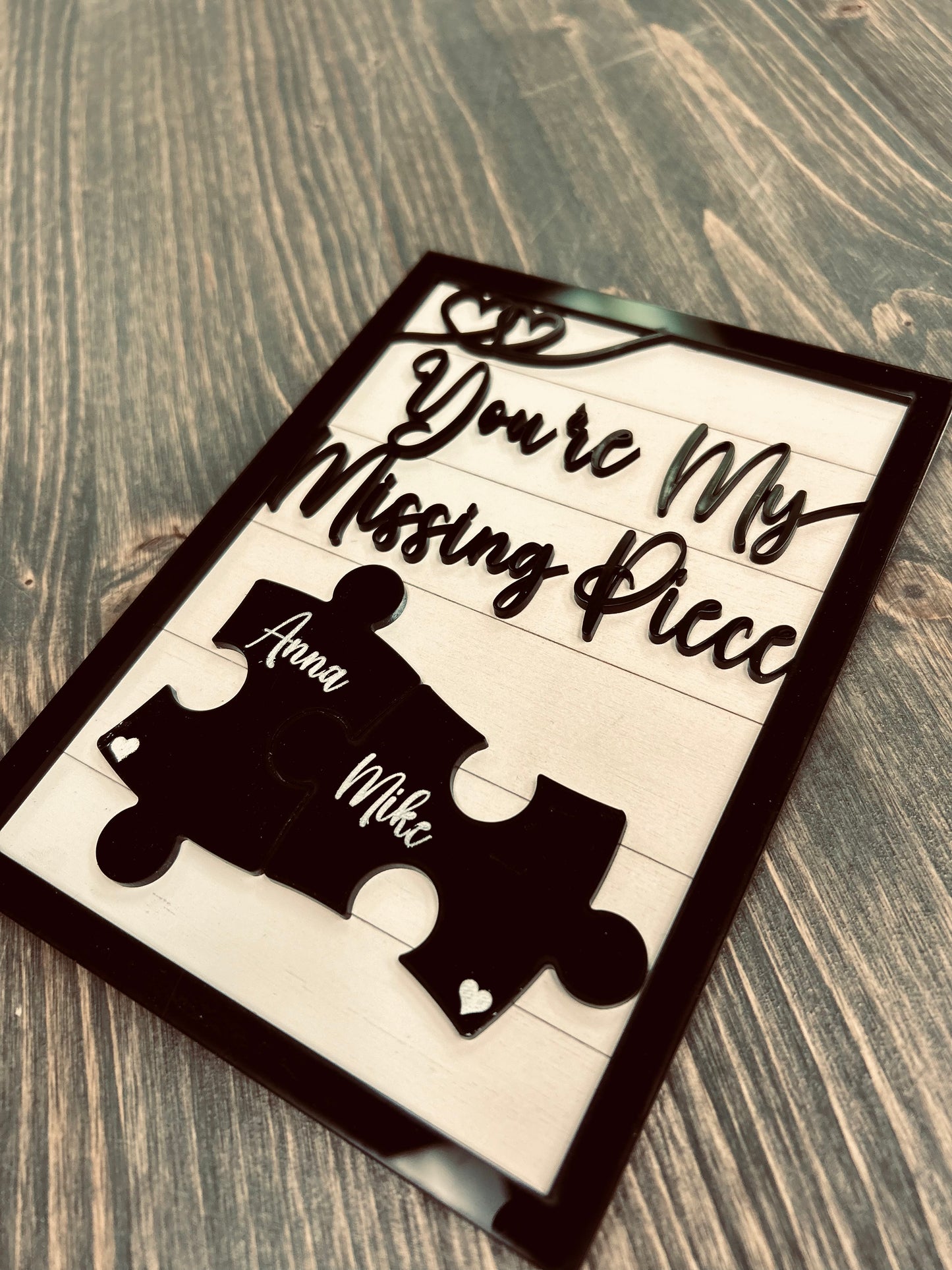 A personalized wooden sign with the phrase "You're My Missing Piece" in elegant black cursive font. Two black puzzle pieces, engraved with the names "Anna" and "Mike," fit together at the center, symbolizing love and connection. The sign is framed in black with small heart details and is displayed on a rustic wooden surface. A meaningful gift idea for couples, anniversaries, or Valentine's Day.