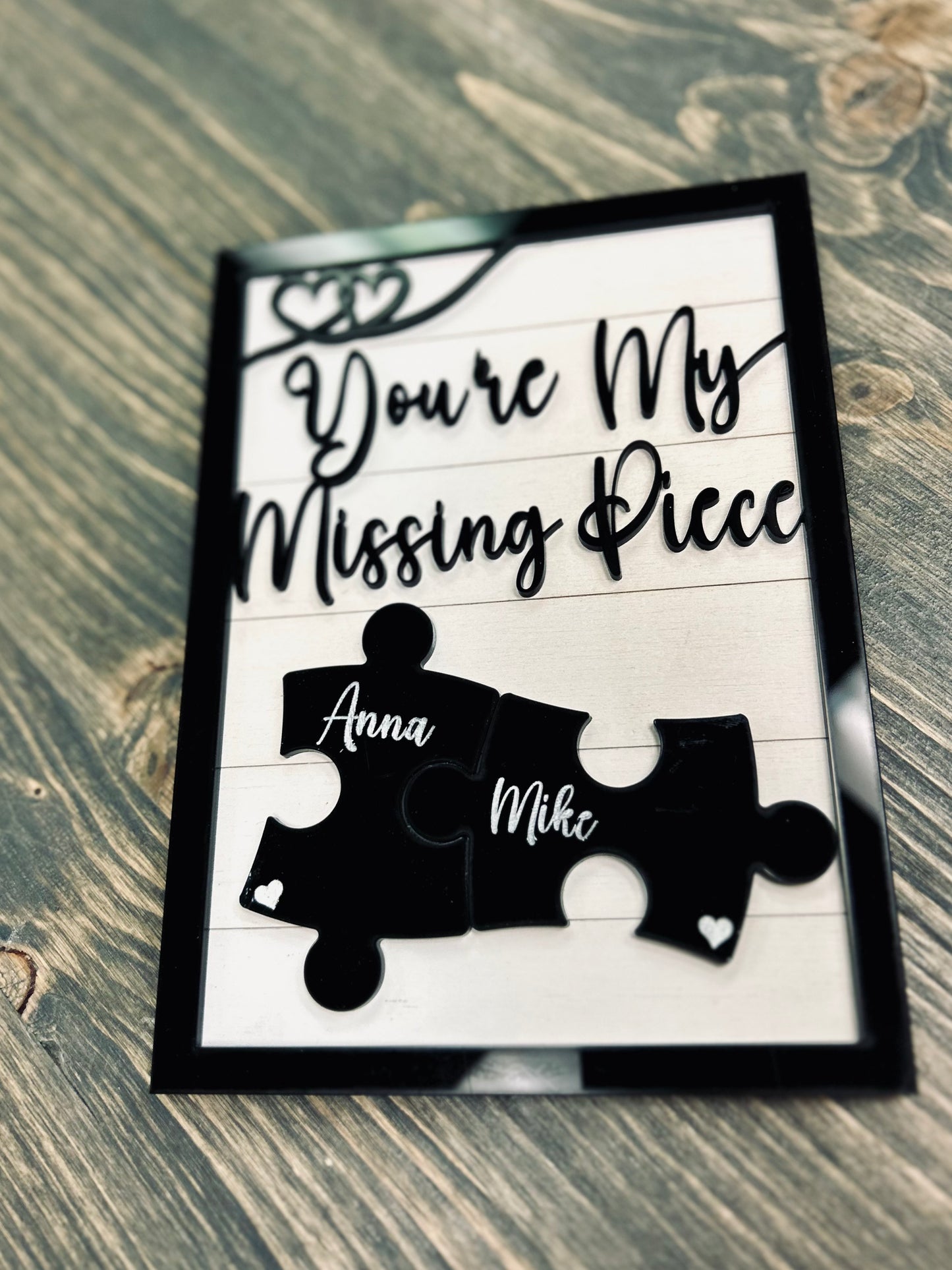 A close-up of a custom wooden sign with the phrase "You're My Missing Piece" in stylish black cursive font. Two black puzzle pieces, engraved with the names "Anna" and "Mike," interlock at the center, symbolizing love and connection. The sign has a black border with heart-shaped accents and rests on a rustic wooden surface. A perfect personalized gift for couples, anniversaries, or Valentine's Day.