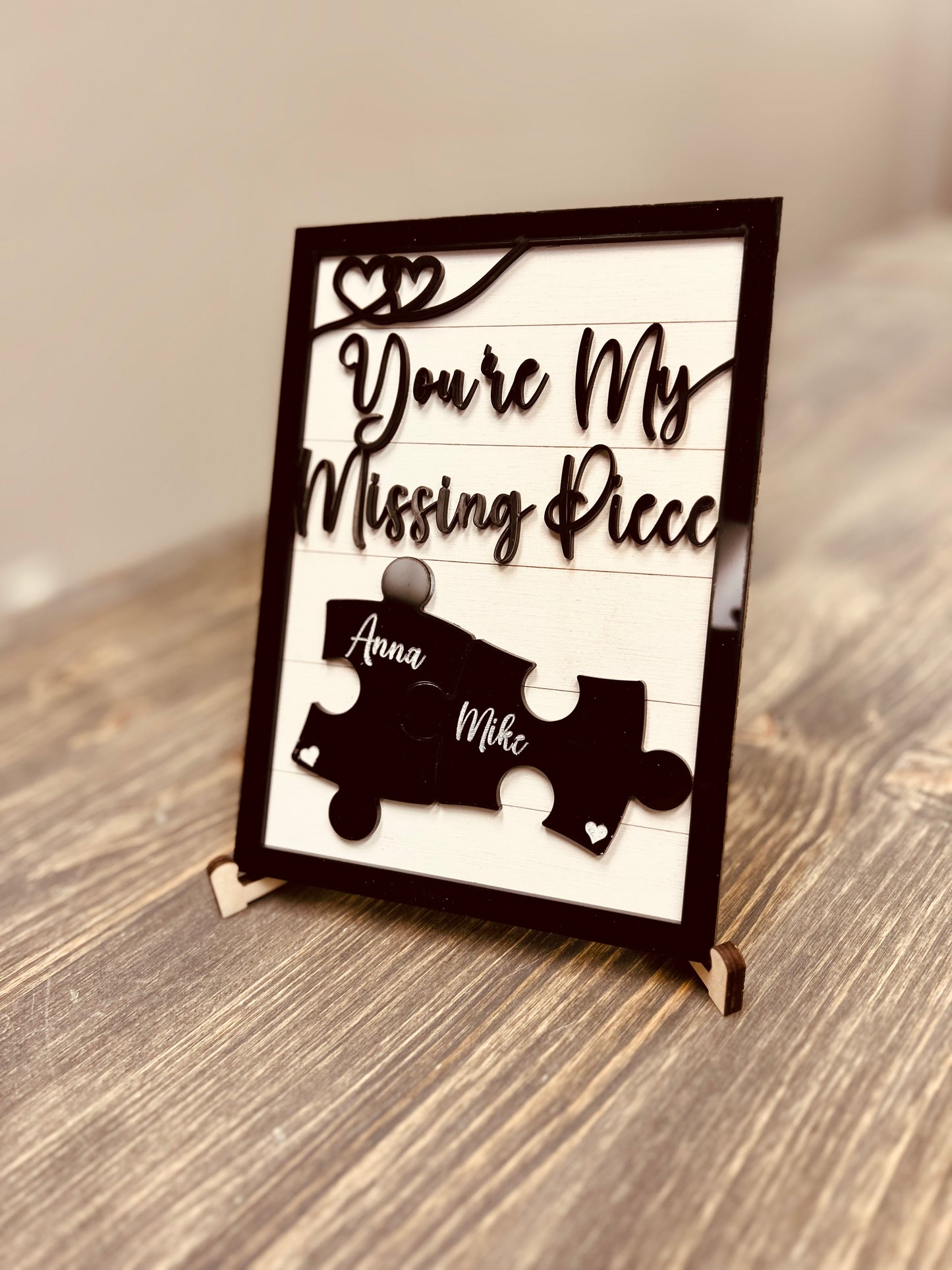 A personalized wooden sign with the phrase "You're My Missing Piece" in elegant cursive font, featuring two interlocking black puzzle pieces engraved with the names "Anna" and "Mike." The sign has a black frame, heart accents, and is displayed on a wooden stand atop a rustic wooden surface. Ideal for romantic gifts, anniversaries, or Valentine's Day.