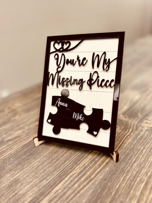 A personalized wooden sign with the phrase "You're My Missing Piece" in elegant cursive font, featuring two interlocking black puzzle pieces engraved with the names "Anna" and "Mike." The sign has a black frame, heart accents, and is displayed on a wooden stand atop a rustic wooden surface. Ideal for romantic gifts, anniversaries, or Valentine's Day.