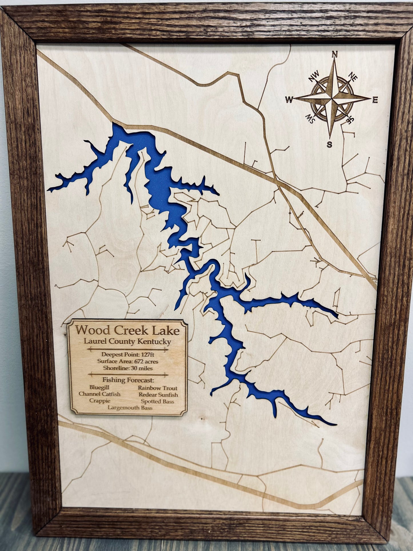 A framed wooden topographic map of Wood Creek Lake in Laurel County, Kentucky, featuring laser-engraved roads and contour lines. The lake is depicted in a deep blue color with an engraved compass rose in the upper right corner. A wooden plaque in the lower left provides lake details, and common fish species such as bluegill, largemouth bass, and rainbow trout. A rustic and decorative piece, perfect for lake enthusiasts and outdoor lovers.