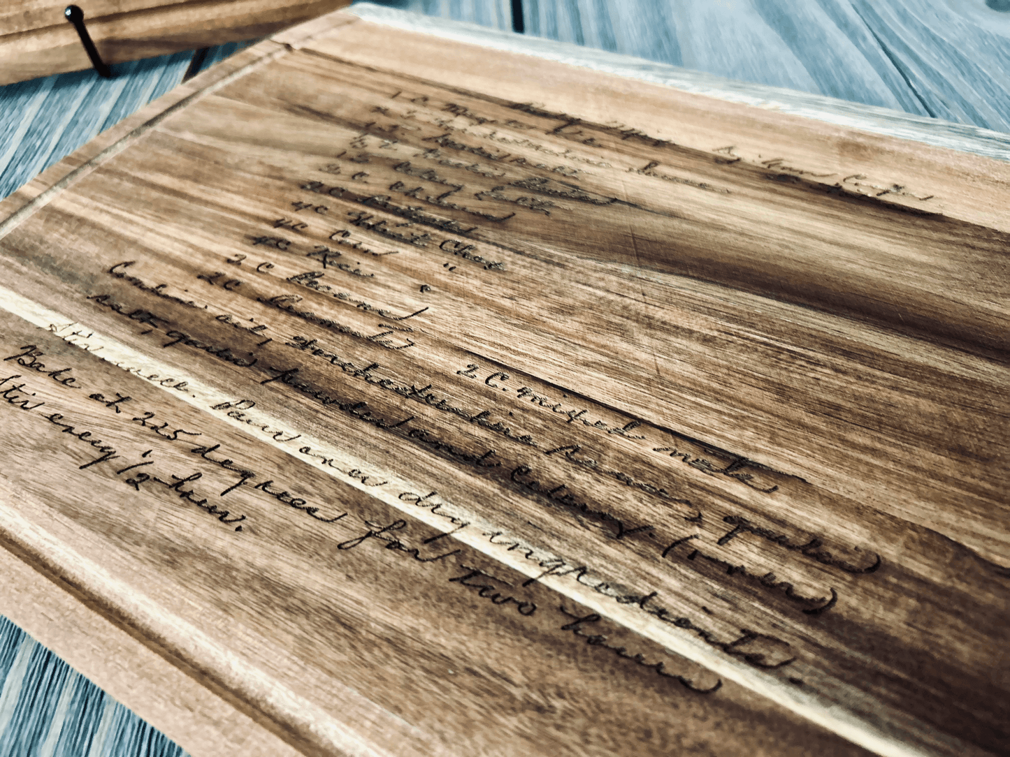 A wooden cutting board with a handwritten engraved recipe, featuring elegant cursive writing. The wood grain and darkened text create a rustic aesthetic. A pen rests in the background on a wooden surface.