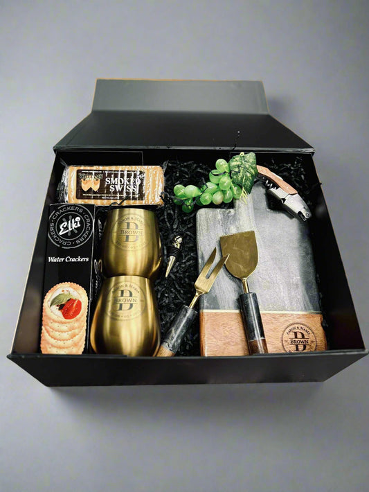 Gift set with gold cups, cheese board, and tools in a black box on a wooden surface.