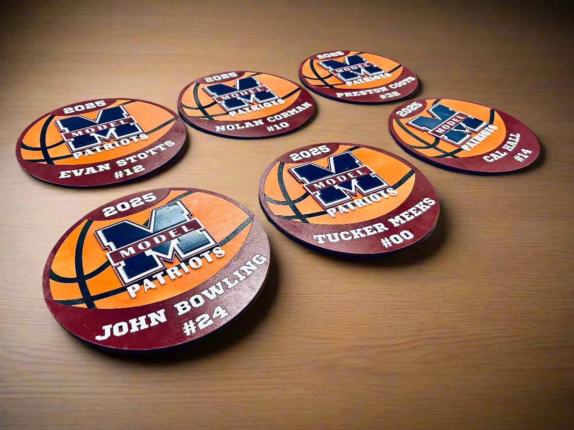 Custom basketball-themed name plaques for the Model Patriots team, featuring an orange basketball design with the team’s 'M' logo at the center. Each plaque is personalized with a player's name, jersey number, and the year '2025' in bold white lettering. The plaques have a circular shape with a maroon border, placed on a wooden surface