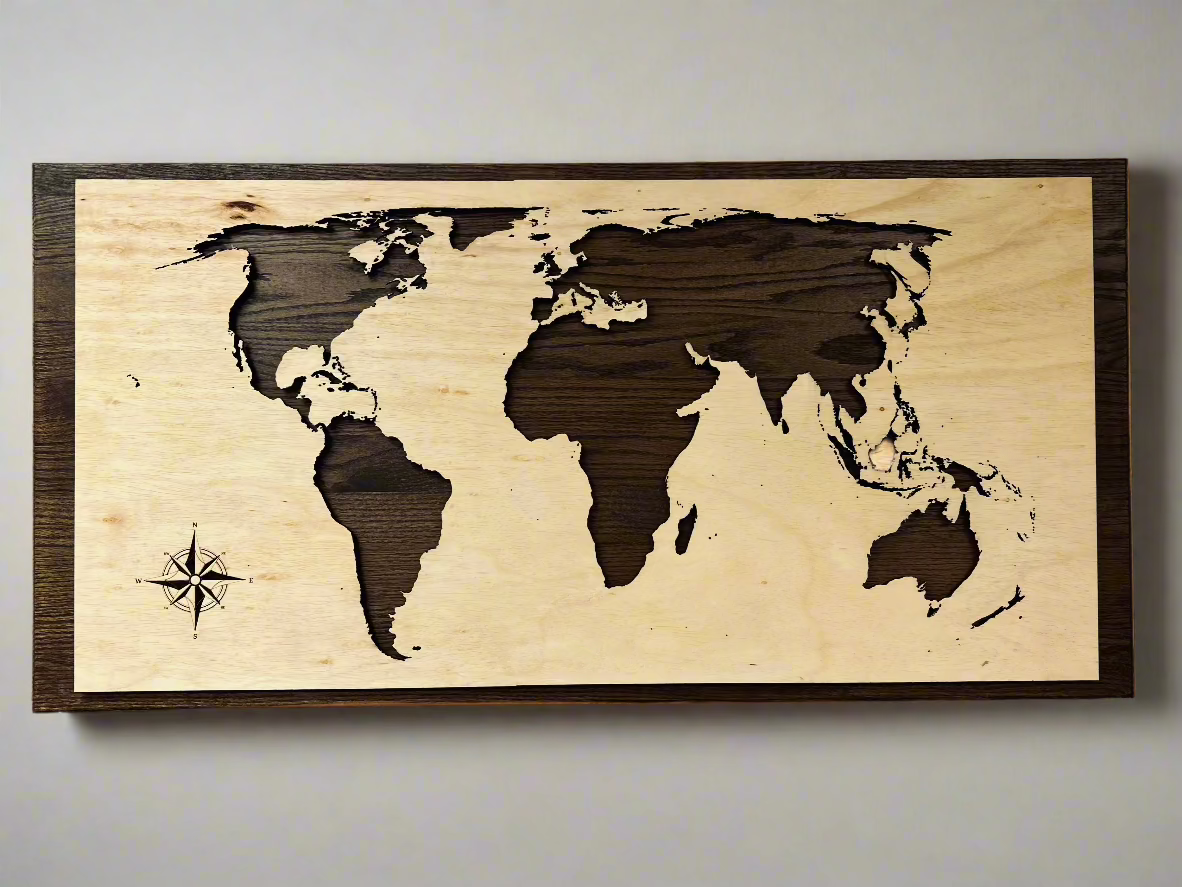 A wooden world map wall art featuring a laser-cut design with light-colored wood for the continents and dark wood for the background. A detailed compass rose is engraved in the lower-left corner. The artwork is framed with a dark wooden border and mounted on a neutral-colored wall.
